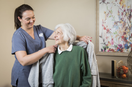 A nurse helps an assisted living resident put her jacket on while smiling at each other