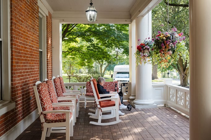 A covered porch with chairs and rocking chairs is shown. A senior woman is sitting in one of the rocking chairs relaxing.