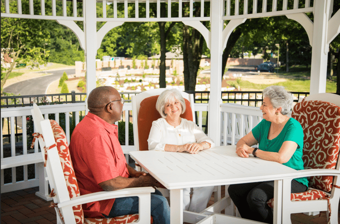 A group of three seniors sit around a patio table under a gazebo smiling at each other.